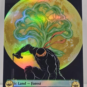Op de afbeelding: Een holografische ruilkaart met een in het zwart geklede ninja met een oranje masker die hurkt voor een grote gele maan en een groen boomsilhouet. De tekst bovenaan luidt Forest en onderaan staat Basic Land - Forest op een zwarte achtergrond.