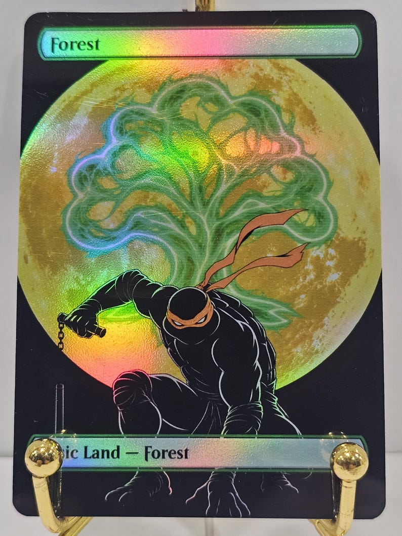 Op de afbeelding: Een holografische ruilkaart met een in het zwart geklede ninja met een oranje masker die hurkt voor een grote gele maan en een groen boomsilhouet. De tekst bovenaan luidt Forest en onderaan staat Basic Land - Forest op een zwarte achtergrond.