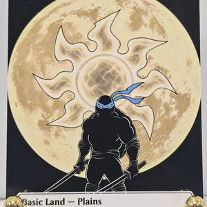 Op de afbeelding: Een rechthoekige ruilkaart met de titel Plains, voorzien van een zwart silhouet van een schildpadfiguur met een blauw masker en twee zwaarden. Achter het personage staat een groot, lichtgeel zonicoon tegen een zwarte achtergrond.