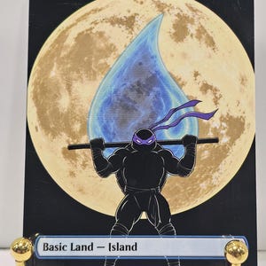 Op de afbeelding: Een rechthoekige ruilkaart met de titel Island, voorzien van een grote gele maan op de achtergrond en een blauw waterdruppelpictogram. Op de voorgrond houdt een zwart silhouet van een schildpad met een paars masker een staf op de schouders.