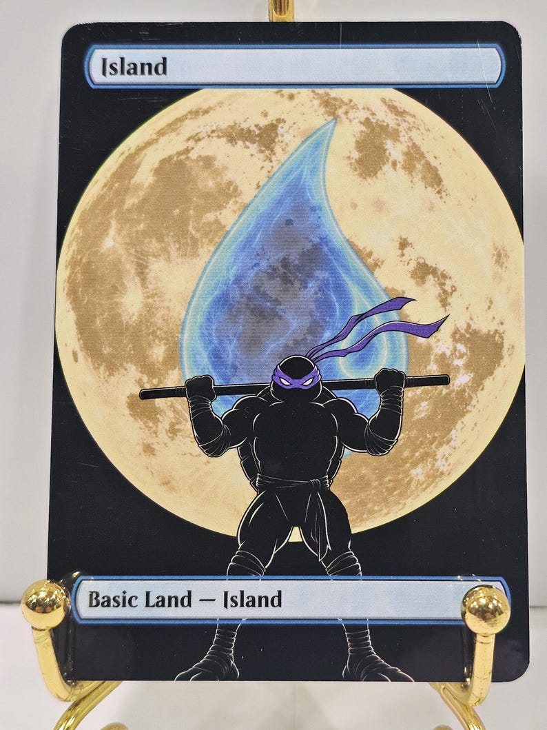Op de afbeelding: Een rechthoekige ruilkaart met de titel Island, voorzien van een grote gele maan op de achtergrond en een blauw waterdruppelpictogram. Op de voorgrond houdt een zwart silhouet van een schildpad met een paars masker een staf op de schouders.
