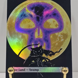 Op de afbeelding: Een rechthoekige ruilkaart met een zwarte rand en een holografische afwerking. De tekst bovenaan luidt Swamp en de tekst onderaan luidt Basic Land - Swamp. Een grote gele maan bevindt zich achter een paars schedelpictogram en een zwart silhouet.