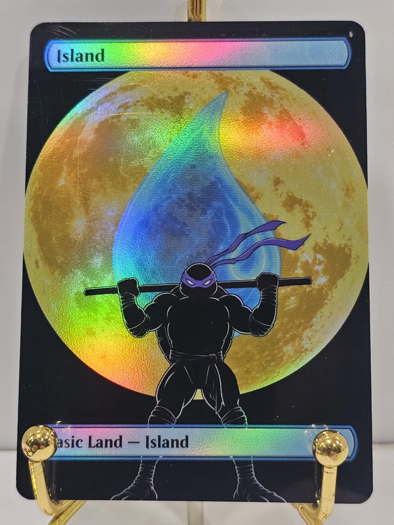 Op de afbeelding: Een rechthoekige ruilkaart met een holografische afwerking met een grote gele maan en een blauw waterdruppelpictogram. Een zwart silhouet van een ninja met een paars masker houdt een staf vast. De tekst Island staat aan de boven- en onderkant.