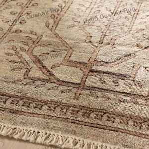 May include: Close-up of a beige and brown patterned rug with a fringe. The rug features a geometric design with tree-like shapes and a border. The text "Hello Oushak Rug" is visible in the image.