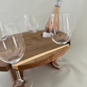 May include: A wooden wine picnic table with a bottle of rosé and two wine glasses. The table has a circular design with a light wood accent. The wine glasses are clear and elegant, and the bottle is partially visible.