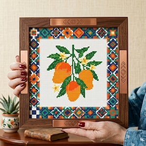 May include: A square cross-stitch embroidery featuring three orange mangoes with green leaves and small yellow flowers, framed in a dark wood border with copper-colored metal accents on each side, each engraved with geometric diamond patterns.