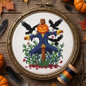 May include: A cross-stitch embroidery in a round bronze-coloured frame featuring a scarecrow with a pumpkin head wearing a blue coat, surrounded by three black crows and green foliage on a white fabric background, with a spool of thread and scissors.