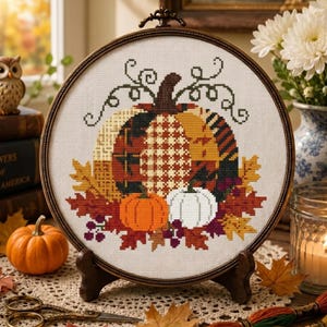 May include: A circular embroidery hoop containing a cross-stitch design of a large central pumpkin with a plaid pattern, flanked by a smaller orange pumpkin and a white pumpkin, surrounded by autumn leaves, berries, and swirling vine details.