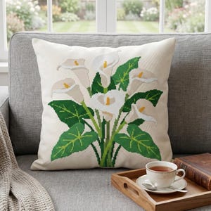 May include: A square throw pillow with a cream-colored fabric base featuring a cross-stitch pattern of white calla lilies with yellow centers and green leaves. The design is centered on the front of the pillow, showcasing a detailed floral motif.