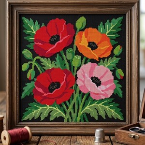 May include: A framed cross-stitch embroidery featuring two red poppies, one orange poppy, and one pink poppy with green leaves and buds on a black background. The artwork is enclosed in a square wooden frame with a visible wood grain texture.