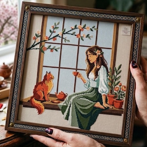 May include: A framed cross-stitch embroidery depicting a person with long brown hair sitting on a windowsill next to a fluffy orange cat. The scene includes a small teapot, a teacup, potted flowers, and a flowering branch against a window grid.
