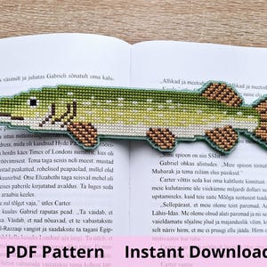 May include: A cross-stitch bookmark shaped like a northern pike fish. The fish features a green and white speckled body with brown patterned fins and tail. The bookmark is placed horizontally across the open pages of a printed book.