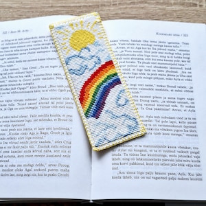 May include: A colorful, hand-stitched bookmark with a rainbow, sun, and clouds design. The bookmark is made of fabric and is placed on top of an open book.