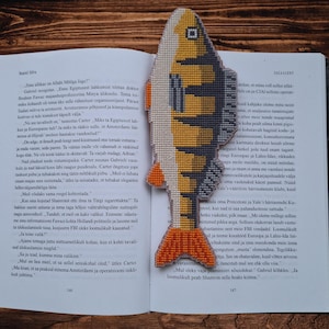 May include: A handmade fish-shaped bookmark made of cross-stitch fabric. The bookmark features a yellow, gray, and orange color scheme, with the fish design placed over an open book. The bookmark is a unique accessory for book lovers.