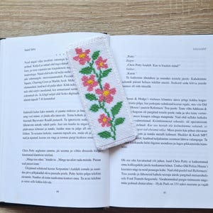 May include: A white bookmark with a floral design in pink and yellow. The bookmark is rectangular and features a repeating pattern of flowers and green leaves. The bookmark is placed on an open book.
