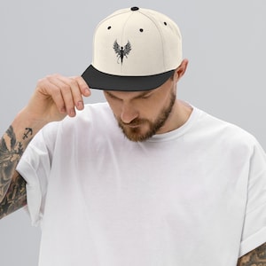 Puede incluir: Gorra de béisbol de ala plana color crema con visera negra y un gráfico negro de una figura alada. La gorra tiene un ajuste estructurado y un cierre a presión. La gorra es usada por una persona.