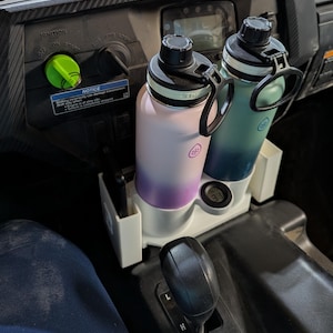 Puede incluir: Un portavasos de plástico blanco con dos botellas de agua aisladas. Una botella es rosa y morada, la otra es turquesa y azul. El soporte también tiene un pequeño compartimento.