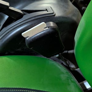 Könnte beinhalten: Nahaufnahme einer schwarzen Kunststoffkomponente mit einem weißen Hebel, wahrscheinlich Teil des Fahrzeuginnenraums. Das Bild zeigt auch einen grün-schwarzen Sitz, was auf Fahrzeug- oder Maschinenteile hindeutet.