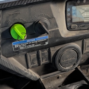 May include: Close-up of a Kawasaki UTV dashboard. Features a green ignition key, various switches, and a digital display. A "NOTICE" sticker provides operational instructions. The dashboard is dark gray with a textured finish.