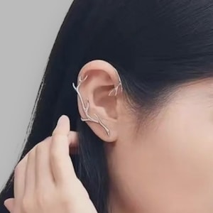 May include: A silver ear cuff shaped like a branch, worn on the ear. The jewellery curves around the ear, with the branch-like design extending along the outer ear. The cuff is a unique accessory.