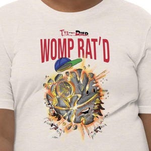 May include: Off-white t-shirt with the text "Tye-Died WOMP RAT'D" in red. The graphic features a cartoon explosion with a baseball cap and a detailed illustration. The shirt is made of a soft material.