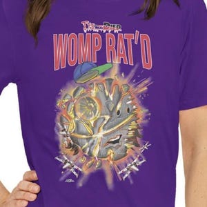 May include: Purple t-shirt with the text "Womp Rat'd" in red and white. The graphic features a cartoon-style design with a baseball cap and a planet-like image. The shirt is short-sleeved.