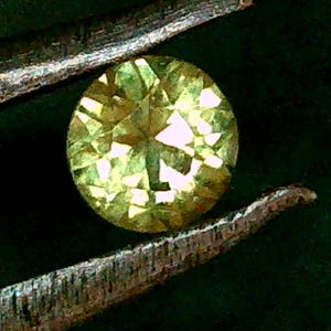 Puede incluir: Una gema redonda y facetada, de color verde claro, sujeta por unas pinzas metálicas. La gema tiene un corte brillante, reflejando la luz. El fondo es verde oscuro, creando contraste. Las pinzas están texturizadas y desgastadas.