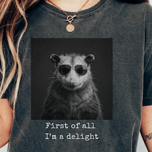 May include: Dark grey t-shirt featuring a black and white image of an opossum wearing sunglasses. The image is in a square and has the text "First of all I'm a delight" below it. The word "PEPPER" is printed in the bottom left corner.