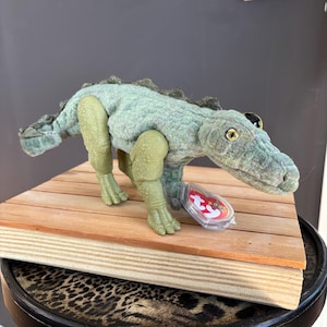 May include: A light green plush alligator toy with a textured, bumpy fabric surface, a long tail, and four legs. The stuffed animal features a prominent snout, yellow and black eyes, and a heart-shaped red and white Ty tag attached to its front leg.