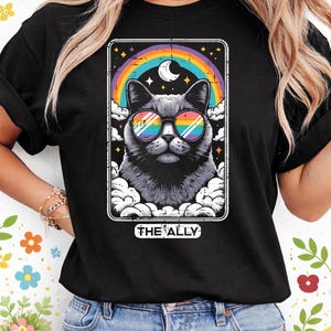 May include: Black t-shirt with a graphic of a cat wearing rainbow sunglasses, set against a rainbow and starry night sky. The cat is framed by clouds, with the text "THE ALLY" below. A vibrant and colourful graphic tee.