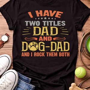 May include: Black t-shirt with the text "I HAVE TWO TITLES DAD AND DOG-DAD AND I ROCK THEM BOTH" in orange, grey, and yellow. The design includes a paw print. The shirt is displayed with jeans, trainers, and a watch.