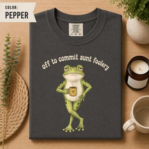 May include: A pepper-coloured t-shirt featuring a graphic of a green frog holding a small yellow mug with a heart icon. Above the frog, the text reads, off to commit aunt foolery. The shirt has a crew neckline and a Comfort Colors brand label.