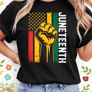May include: Black t-shirt featuring a graphic of a raised fist within a flag design with red, yellow, green, and stars. The word "JUNETEENTH" is printed vertically in white. The shirt is worn by a person.