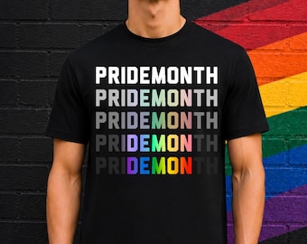 Könnte beinhalten: Schwarzes T-Shirt mit dem Text "PRIDEMONTH" in einem gestapelten Design. Das Wort ist in Weiß, mit dem unteren Wort in Regenbogenfarben. Der Hintergrund ist eine schwarze Backsteinmauer mit einem Regenbogen darauf.