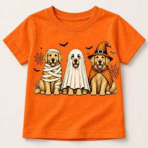 May include: A bright orange short-sleeved t-shirt featuring a graphic of three golden retriever dogs dressed for Halloween. One dog is wrapped like a mummy, one wears a white ghost sheet, and the third wears a witch hat and orange cape.