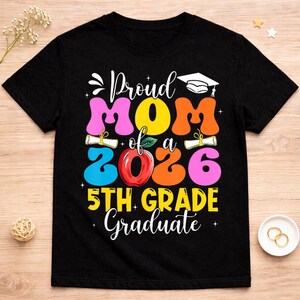 以下が含まれることがあります： 「Proud Mom of a 2026 5th Grade Graduate」と書かれたカラフルなグラフィックが特徴の黒いTシャツ。卒業帽子、巻物、赤いリンゴのデザインが含まれています。テキストはピンク、オレンジ、黄色、青など様々な色で表示されています。