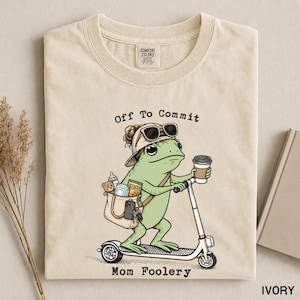 Könnte beinhalten: Ein elfenbeinfarbenes T-Shirt mit einer Grafik eines grünen Frosches mit Baseballkappe und Sonnenbrille, der auf einem Roller fährt und eine Tasche mit Babyflasche, Teddybär, Feuchttüchern, Snacks und Schlüsseln trägt, mit dem Text Off To Commit Mom Foolery.