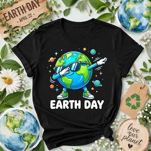 Puede incluir: Camiseta negra con un personaje de la Tierra de dibujos animados con gafas de sol y haciendo un dab. Las palabras "EARTH DAY" están en blanco. Planetas y estrellas rodean la Tierra. Un letrero de madera muestra "EARTH DAY APRIL 22".