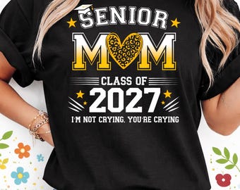 May include: Black t-shirt with the text "SENIOR MOM CLASS OF 2027". The design includes a heart with a leopard print pattern and stars. The phrase "I'M NOT CRYING, YOU'RE CRYING" is printed below the year.