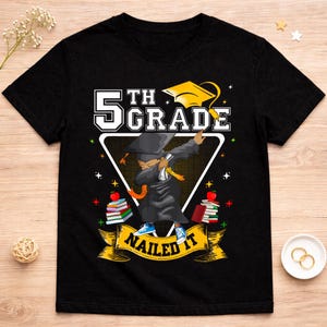 May include: Black t-shirt with a graphic celebrating 5th grade graduation. The design features a student dabbing in a graduation gown and cap, with the words "5th Grade" and "Nailed It" in a banner. Books and apples are also included.