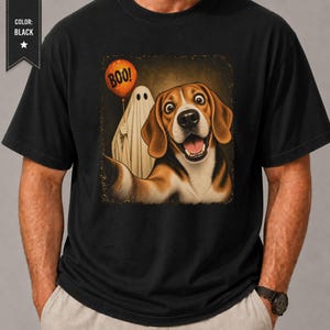 May include: A black short-sleeved t-shirt featuring a graphic print of a beagle dog with a wide-eyed expression and an open mouth. Beside the dog is a white ghost figure holding an orange balloon with the word BOO printed in black letters.