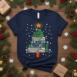 Puede incluir: Camiseta azul marino con un diseño de árbol de Navidad hecho con componentes de ordenador, incluyendo teclados, placas base y ratones. Una estrella dorada corona el árbol, con copos de nieve cayendo. Decoraciones navideñas.