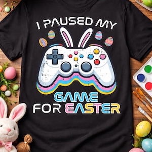Op de afbeelding: Zwart t-shirt met een wit videogamecontroller-ontwerp. De controller heeft konijnenoren en paasei-details. De tekst op het shirt luidt "I PAUSED MY GAME FOR EASTER". Kleurrijk ontwerp.