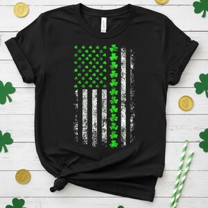 May include: Black t-shirt featuring a distressed American flag design with green shamrocks replacing the stars and stripes. The flag design includes white stripes and green shamrock accents, perfect for St. Patrick's Day.
