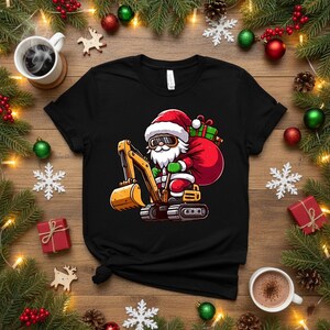 Peut inclure: T-shirt noir avec un dessin de Père Noël conduisant une excavatrice jaune. Le Père Noël porte un bonnet rouge et une tenue verte, avec un sac de cadeaux. Décoré d'ornements de Noël et de verdure.