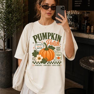 May include: An off-white short-sleeve t-shirt featuring a graphic print of an orange pumpkin with green leaves and flowers. The text reads PUMPKIN PATCH, FRESH FROM THE PATCH, FARM FRESH, LOCALLY GROWN, PUMPKINS, CORN MAZE, and APPLE CIDER.