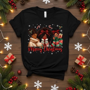May include: Black t-shirt featuring a graphic of three Black women in winter attire, with the text "Merry Christmas" in red script. The women are adorned with festive accessories and surrounded by snowflakes and candy canes.