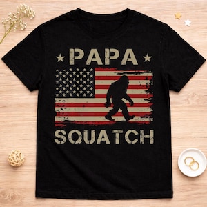 May include: Black t-shirt featuring the text "PAPA SQUATCH" with an American flag design and a silhouette of Bigfoot. The design has a distressed, vintage look. The shirt is made of a soft material.