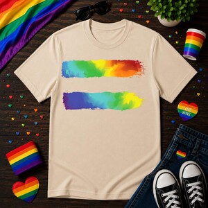 Puede incluir: Una camiseta beige con dos pinceladas horizontales de colores del arcoíris. La imagen incluye una bandera arcoíris, gafas de sol, una planta pequeña, una taza a rayas arcoíris y unos vaqueros con zapatillas negras.
