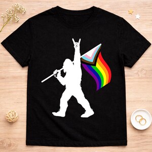 May include: Black t-shirt featuring a white silhouette of Bigfoot holding a flag with a rainbow and transgender pride flag design. The Bigfoot is making a rock-on hand gesture. The flag has a rainbow design.
